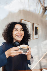 Happy curly hair woman with coffee cup seen through glass at cafe - JOSEF11882