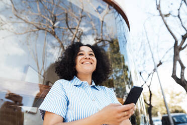 Happy curly hair woman with mobile phone leaning on glass - JOSEF11894