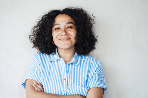 Smiling curly hair woman with arms crossed standing in front of wall - JOSEF11902