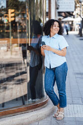 Smiling woman with smart phone leaning on glass at footpath - JOSEF11916