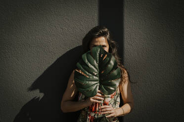 Mature woman covering face with monstera leaf in front of wall - MJRF00747