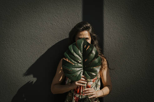 Reife Frau bedeckt ihr Gesicht mit einem Monstera-Blatt vor einer Wand - MJRF00747