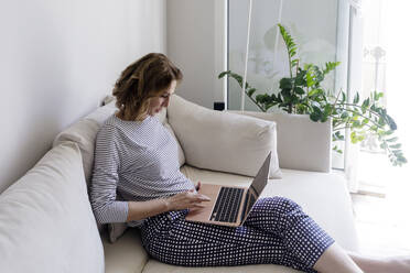 Mature woman using laptop sitting on sofa at home - RFTF00267