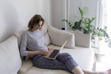 Smiling woman with laptop sitting on couch at home - RFTF00268