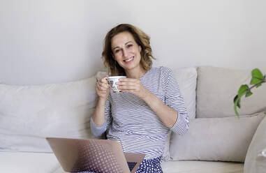 Lächelnde Frau mit Laptop und Kaffeetasse, die zu Hause auf der Couch sitzt - RFTF00275