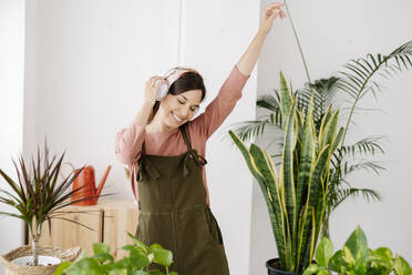 Smiling woman listening music through headphones dancing amidst plants - JCZF01039