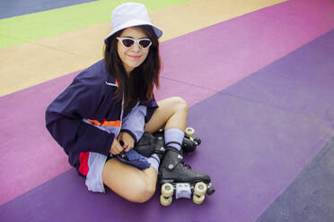 Smiling woman wearing roller skates sitting on multi colored floor - EGCF00011