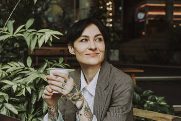 Lächelnde Geschäftsfrau mit kurzen Haaren, die einen Einweg-Kaffeebecher hält und in einem Straßencafé sitzt - OIPF02324