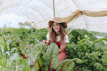 Smiling farm worker with clipboard at greenhouse - MRRF02361