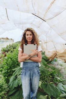 Smiling farm worker with clipboard standing in greenhouse - MRRF02383