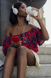 Young woman wearing headphones drinking coffee from disposable cup on steps - PGF01180