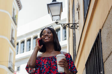 Lächelnde Frau, die einen Einweg-Kaffeebecher hält und mit einem Smartphone an einem Gebäude telefoniert - PGF01182