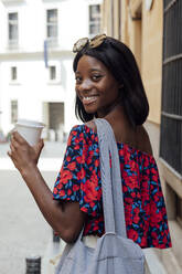 Happy woman wearing shoulder bag and sunglasses holding disposable cup - PGF01183