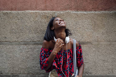 Fröhliche Frau mit Kopfhörern, die vor einer Wand nach oben schaut - PGF01185