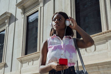 Junge Frau mit Smartphone hört Musik über Kopfhörer an einem sonnigen Tag - PGF01194
