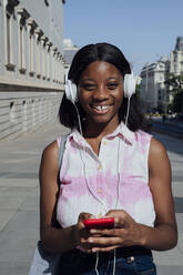 Glückliche Frau mit Mobiltelefon hört Musik über Kopfhörer in der Stadt an einem sonnigen Tag - PGF01196