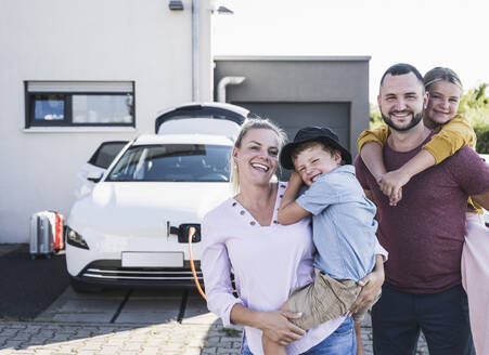 Glückliche Familie steht vor dem Haus der Familie und lädt ein Elektroauto auf - UUF27088