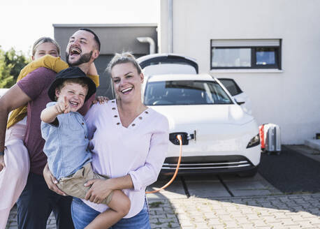 Glückliche Familie vor dem Haus der Familie und dem Elektroauto - UUF27089