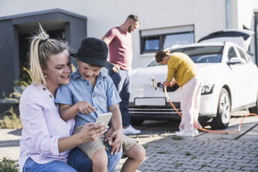 Mutter und Sohn prüfen ihr Smartphone, während Vater und Tochter ihr Elektroauto aufladen - UUF27091