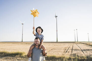 Vater trägt seinen Sohn mit einem Windrad auf den Schultern vor einer Windkraftanlage - UUF27127