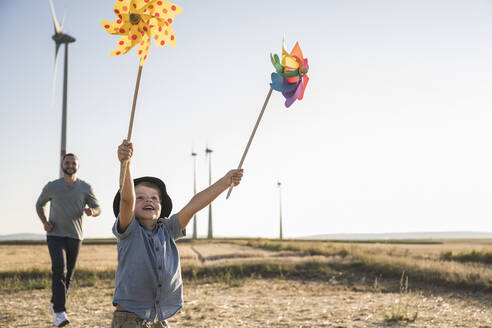 Boy with colorful pinwheels running in windpark with father following - UUF27132