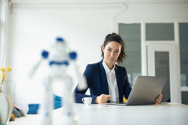 Serious businesswoman sitting with laptop looking at toy robot on table - JOSEF11946