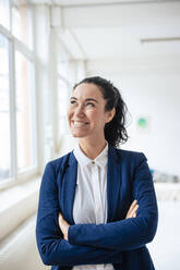 Happy businesswoman standing with arms crossed by window - JOSEF11960