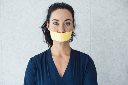 Woman with tape over mouth standing in front of gray wall - JOSEF11979