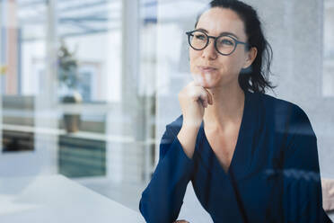 Smiling businesswoman sitting with hand on chin seen through glass - JOSEF11985