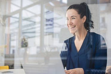 Cheerful businesswoman with laptop seen through glass - JOSEF11988
