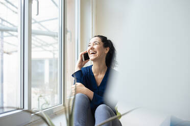Glückliche Geschäftsfrau, die am Fenster sitzend mit ihrem Handy telefoniert - JOSEF11997