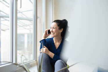 Smiling woman talking on mobile phone sitting by window - JOSEF11998