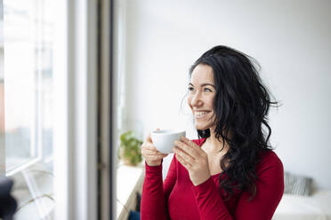 Glückliche Frau hält Kaffeetasse und schaut durch das Fenster zu Hause - JOSEF12010