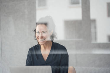 Glückliche Geschäftsfrau mit Laptop vor einer Wand durch Glas gesehen - JOSEF12030