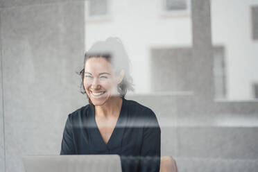 Happy businesswoman with laptop in front of wall seen through glass - JOSEF12030