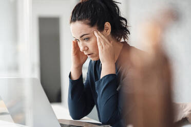 Stressed businesswoman with head in hands looking at laptop on table - JOSEF12032