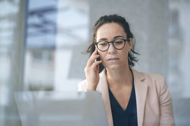 Businesswoman talking on mobile phone sitting with laptop in front of gray wall seen through glass - JOSEF12035