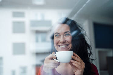 Happy woman holding coffee cup seen through glass - JOSEF12057