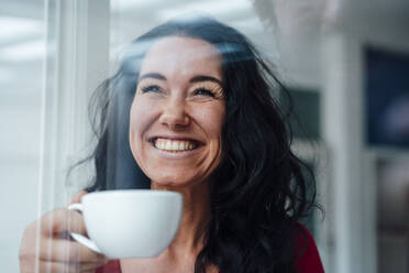 Cheerful woman holding coffee cup seen through glass - JOSEF12059