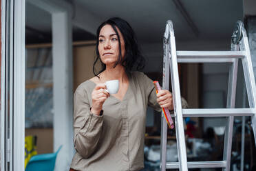 Nachdenkliche Frau mit Kaffeetasse, die zu Hause auf einer Leiter steht - JOSEF12064