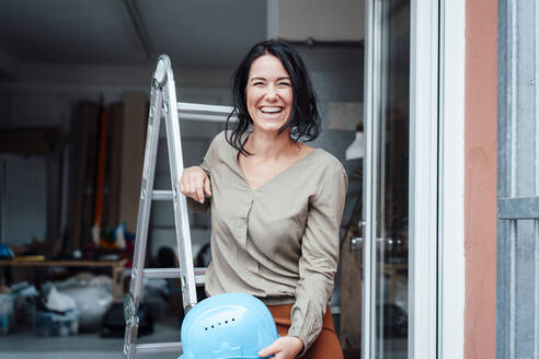 Happy woman holding hardhat leaning on ladder at entrance of home - JOSEF12067