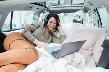 Happy woman wearing headphones watching movie on laptop by lying in car trunk - JOSEF12070