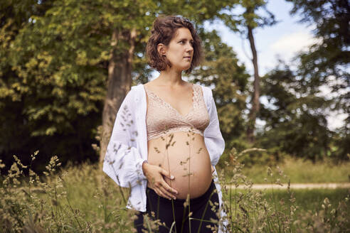 Schwangere Frau steht mit der Hand auf dem Bauch im Gras - JHAF00157