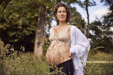 Lächelnde schwangere Frau steht mit der Hand auf der Hüfte im Gras - JHAF00158