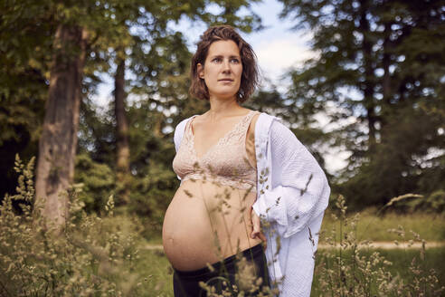 Nachdenkliche schwangere Frau, die mit der Hand auf der Hüfte im Gras steht - JHAF00159