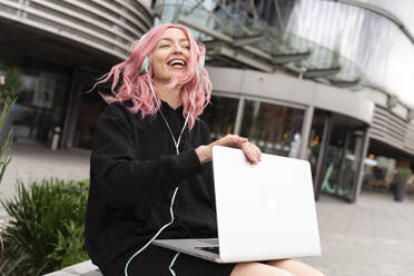 Happy young woman with pink hair listening music through headphones in front of building - TOF00020