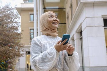 Glückliche junge Frau mit Smartphone vor einem Gebäude stehend - AMWF00295