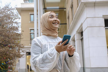 Happy young woman with smart phone standing in front of building - AMWF00295