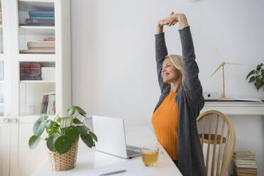 Smiling freelancer stretching arms at desk in home office - SVKF00499