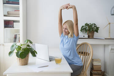 Glückliche blonde Freiberuflerin streckt die Arme am Schreibtisch im Home Office aus - SVKF00504
